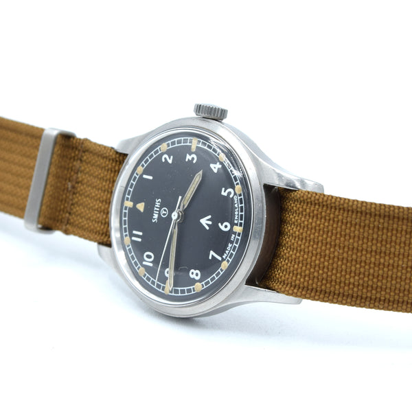A vintage-style wristwatch with a black dial, silver-tone metal case, and a brown canvas strap, photographed on a clean white background.