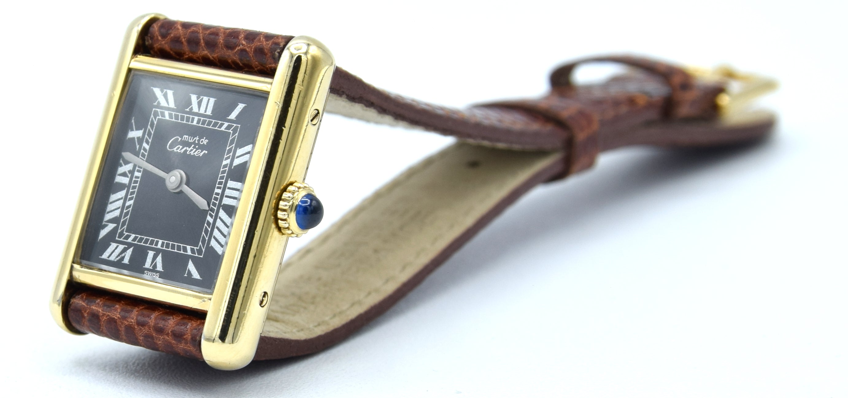 Cartier Tank watch with a gold-tone case, black dial, Roman numeral markers, and a brown leather strap with a blue cabochon accent on the crown.
