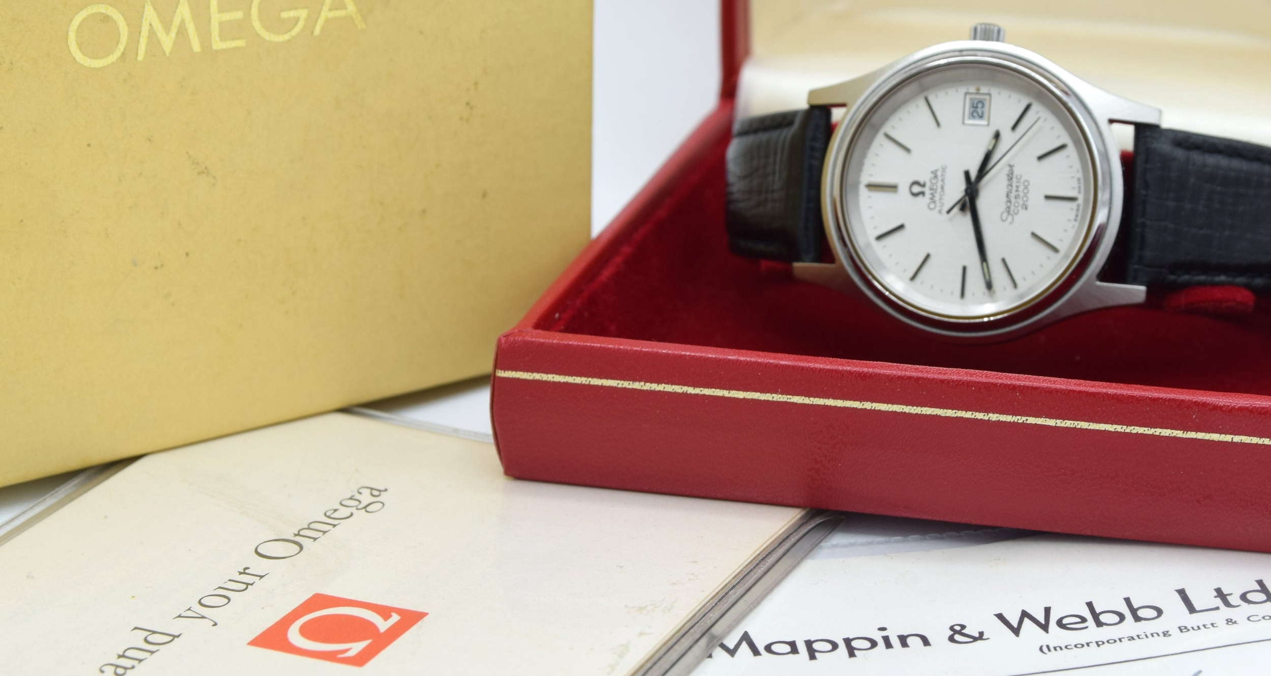 Omega wristwatch displayed in an open red presentation box, with the gold Omega box lid, booklet, and papers visible around it.